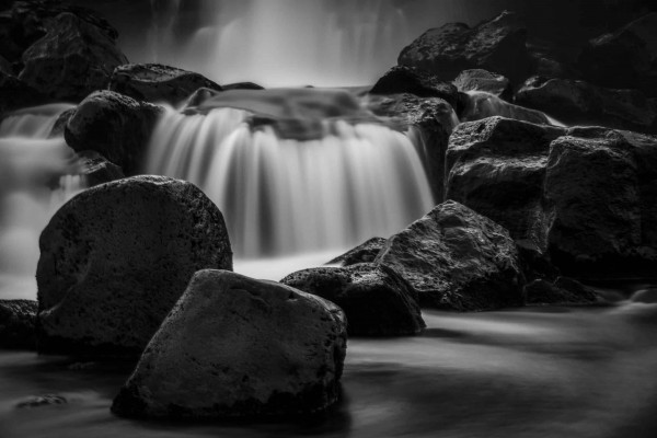 Svartvit bild på ett litet vattenfall bakom några stenar. Fotograf: Susanne Kvarnlöf
